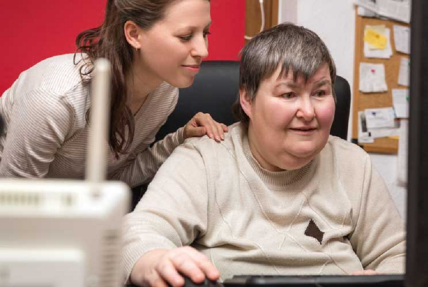 an older woman with a developmental disability sits at a computer with her direct support professional standing behind her
