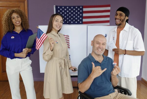 You Uploaded image Generate alt text for this image Copilot A group of four people is standing and sitting in front of a voting booth with an American flag in the background. One person is holding a small American flag, and another person is holding a clipboard. The person in the wheelchair is showing a "thumbs up" gesture and has an "I Voted" sticker on the shirt. Another person is holding a piece of paper, possibly a ballot.