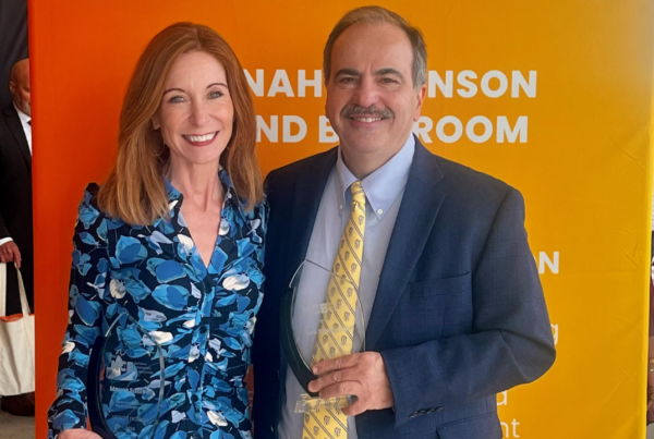 Two individuals are standing in front of an orange backdrop with white text. One person is wearing a blue floral dress, and the other is wearing a suit with a yellow tie. They are each holding a glass award.