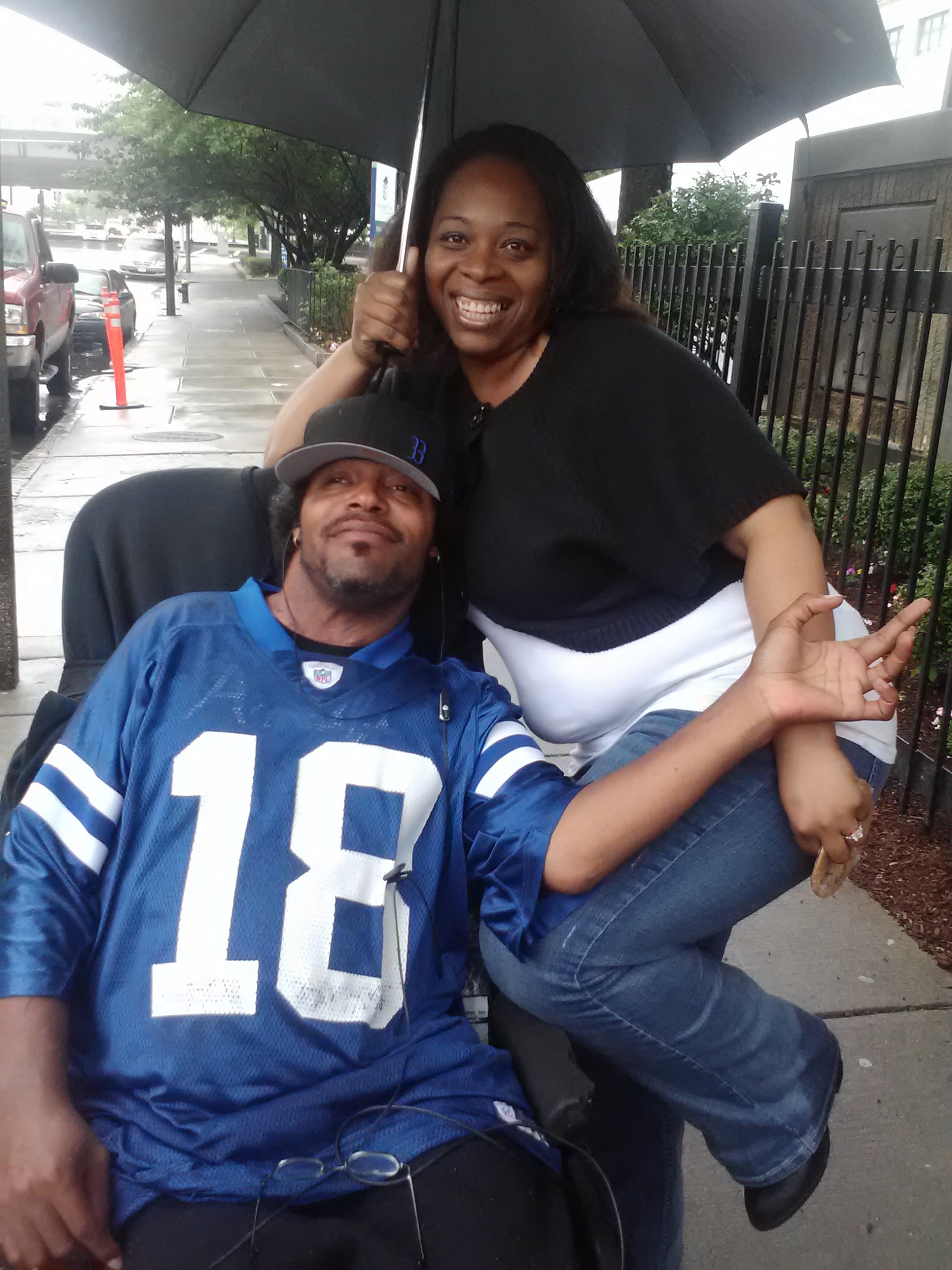 A man in a wheelchair wearing a blue shirt with the number 18 on it, sitting under an umbrella being held by a smiling woman wearing jeans and a black shirt.