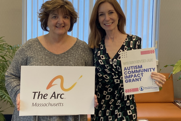 Two women stand side by side. The woman on the left holds a sign that says The Arc of Massachusetts. The woman on the right holds a sign that says We Received An Autism Community Impact Grant.