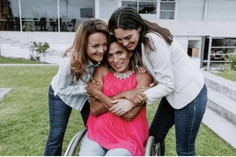 Three women, the one in the middle using a wheelchair, who are hugging and smiling