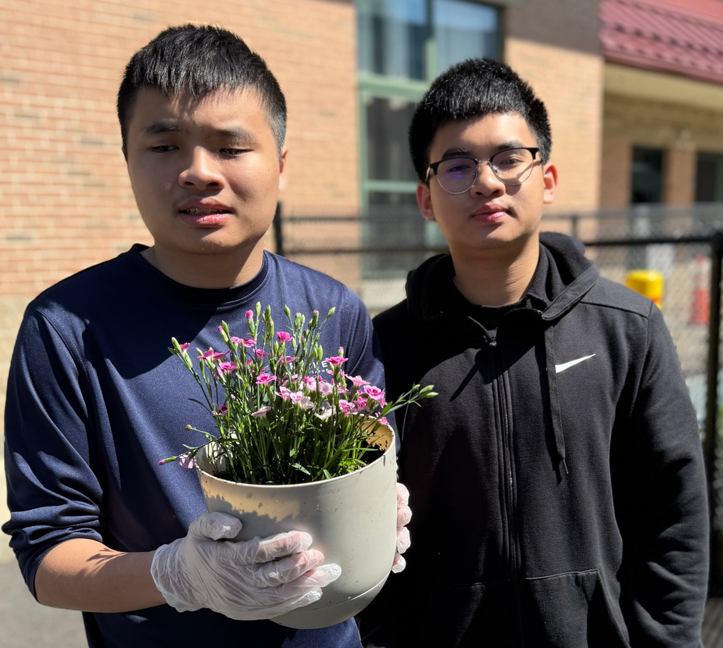 Two young men wearing plastic gloves, with one holding a pot of flowers