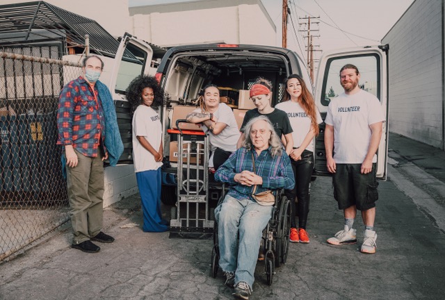 7 people, one of whom uses a wheelchair, standing in front of a truck full of supplies and using a dolly